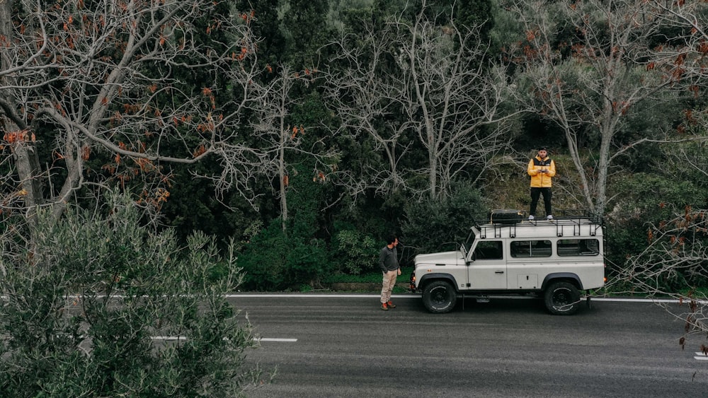 man standing in front of white Land Rover Defender SUV and other man standing on top of it during daytime