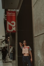 A fashionable person leans against a wall beside a red sign reading 'Retro'. They wear sunglasses, a patterned crop top, and blue pants, exuding a confident and stylish demeanor. Other people are walking along the street in the background.