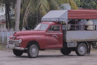 An old, rustic red truck with a covered rear cargo area is parked beside a road. The truck appears weathered and worn, with visible rust patches on its metal surface. Several people are sitting inside the vehicle's open back under a metal canopy, which is also partially rusted. Palm trees and a fence line the background, giving a tropical setting.