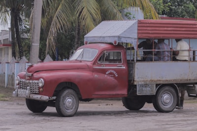 An old, rustic red truck with a covered rear cargo area is parked beside a road. The truck appears weathered and worn, with visible rust patches on its metal surface. Several people are sitting inside the vehicle's open back under a metal canopy, which is also partially rusted. Palm trees and a fence line the background, giving a tropical setting.