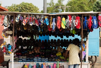 A small market stall is displaying a wide variety of colorful sandals and flip-flops. They are hanging from the roof and also arranged on the counter. A child is seen looking at the footwear.