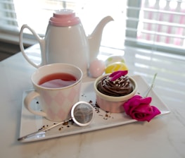 A white teapot with a pink lid sits near a teacup filled with red tea on a white tray. Beside it is a small ramekin with chocolate frosting, topped with petals and sprinkles. A bright pink rose lies next to the dessert. The setup is on a marble surface, near a window with blinds.