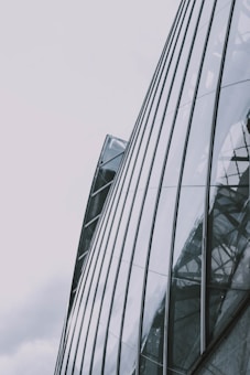 A modern architectural structure consisting of curved glass panels. The reflective surface captures abstract patterns and the sky, creating a sleek and contemporary appearance.