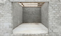 An unfinished brick structure with three visible walls, lacking a front wall and a roof, creating a rectangular opening. The bricks appear grey and uniform, and the ground is covered with a smooth, light-colored material. Wooden beams are seen partially atop the structure, suggesting ongoing construction work.