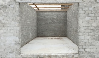 An unfinished brick structure with three visible walls, lacking a front wall and a roof, creating a rectangular opening. The bricks appear grey and uniform, and the ground is covered with a smooth, light-colored material. Wooden beams are seen partially atop the structure, suggesting ongoing construction work.