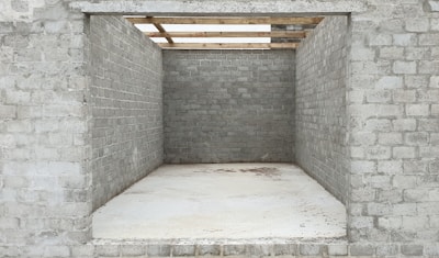 An unfinished brick structure with three visible walls, lacking a front wall and a roof, creating a rectangular opening. The bricks appear grey and uniform, and the ground is covered with a smooth, light-colored material. Wooden beams are seen partially atop the structure, suggesting ongoing construction work.