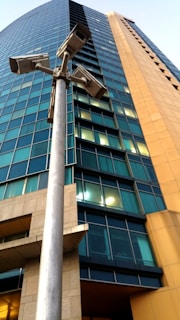 A tall modern building with a sleek facade of blue-tinted reflective windows. A prominent metal pole in the foreground has multiple security cameras attached, suggesting surveillance. The building features alternating sections of glass and concrete with an architectural design that combines sharp angles with smooth curves.