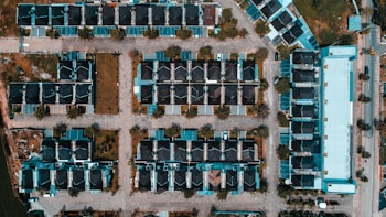 An aerial view of a suburban housing complex with rows of houses arranged in a grid-like pattern. The houses feature dark roofs and light-colored walls. Streets with trees are laid out between the houses, and a large rectangular building is visible on the right side. The landscape includes patches of greenery and several parked vehicles.