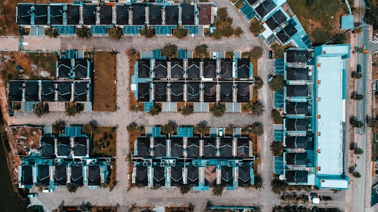 An aerial view of a suburban housing complex with rows of houses arranged in a grid-like pattern. The houses feature dark roofs and light-colored walls. Streets with trees are laid out between the houses, and a large rectangular building is visible on the right side. The landscape includes patches of greenery and several parked vehicles.