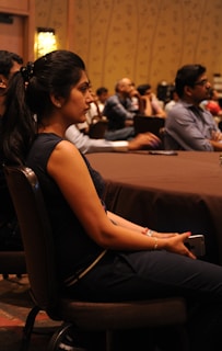 A woman with long dark hair, wearing a dark top and pants, sits attentively in a chair with a smartphone in her hand. She faces forward in what appears to be a conference or seminar setting. The background shows other attendees seated at a long table in a room with dim lighting and patterned walls.