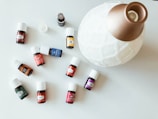 A collection of small essential oil bottles with white caps is spread out on a light surface. Each bottle has a distinct label with various colors, including red, blue, yellow, green, and orange. A large, white, ceramic diffuser with a golden top is positioned next to the bottles.