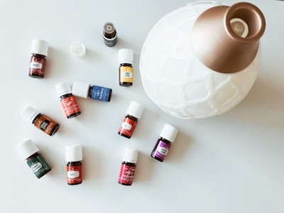 A collection of small essential oil bottles with white caps is spread out on a light surface. Each bottle has a distinct label with various colors, including red, blue, yellow, green, and orange. A large, white, ceramic diffuser with a golden top is positioned next to the bottles.