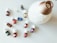 A collection of small essential oil bottles with white caps is spread out on a light surface. Each bottle has a distinct label with various colors, including red, blue, yellow, green, and orange. A large, white, ceramic diffuser with a golden top is positioned next to the bottles.