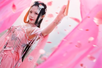 A woman dressed in an elaborate, intricate pink traditional outfit with floral patterns and delicate accessories. She is surrounded by flowing pink fabric and petals, creating a dynamic and ethereal scene. Her expression is serene and confident.