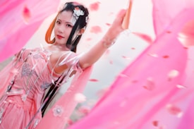 A woman dressed in an elaborate, intricate pink traditional outfit with floral patterns and delicate accessories. She is surrounded by flowing pink fabric and petals, creating a dynamic and ethereal scene. Her expression is serene and confident.