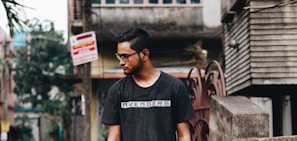 A young man with short hair and glasses stands outdoors in an urban setting. He is wearing a dark shirt with 'Avengers' written on it. The background features a mix of residential and commercial buildings, with various signs and foliage.