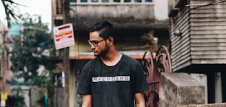 A young man with short hair and glasses stands outdoors in an urban setting. He is wearing a dark shirt with 'Avengers' written on it. The background features a mix of residential and commercial buildings, with various signs and foliage.