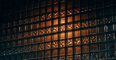 A wall composed of translucent glass blocks illuminated with a warm orange light, creating a pattern of reflections and shadows. The surface texture is intriguing with complex play of light and dark areas.