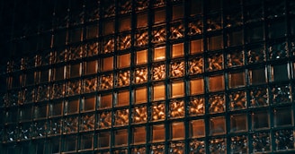 A wall composed of translucent glass blocks illuminated with a warm orange light, creating a pattern of reflections and shadows. The surface texture is intriguing with complex play of light and dark areas.