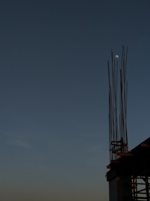 The image features a construction site with steel rods extending upwards against a clear evening sky. The moon is visible in the background, providing a serene and calm atmosphere.