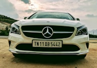 A front view of a white Mercedes-Benz car with a clear focus on the grille and emblem. The car is parked on a sandy surface with a cloudy sky backdrop, flanked by a mountainous landscape.