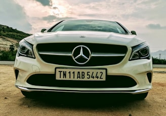 A front view of a white Mercedes-Benz car with a clear focus on the grille and emblem. The car is parked on a sandy surface with a cloudy sky backdrop, flanked by a mountainous landscape.