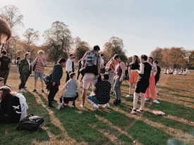 A group of people gathered in a park, interacting and enjoying the outdoors. The area is grassy with some people standing and others sitting on the ground. Trees with autumn foliage are visible in the background, and the scene suggests a casual, social atmosphere.