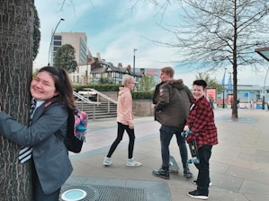 A group of young people are enjoying themselves in an urban setting. One person is joyfully hugging a tree while others are walking nearby. The scene includes modern buildings, trees, and a clear sky, indicating a city environment.