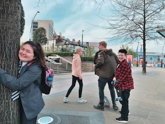 A group of young people are enjoying themselves in an urban setting. One person is joyfully hugging a tree while others are walking nearby. The scene includes modern buildings, trees, and a clear sky, indicating a city environment.