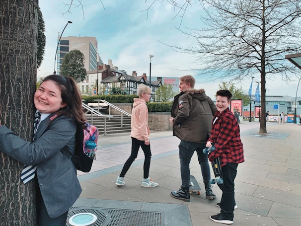 A group of young people are enjoying themselves in an urban setting. One person is joyfully hugging a tree while others are walking nearby. The scene includes modern buildings, trees, and a clear sky, indicating a city environment.