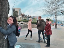 A group of young people are enjoying themselves in an urban setting. One person is joyfully hugging a tree while others are walking nearby. The scene includes modern buildings, trees, and a clear sky, indicating a city environment.