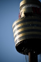A tall, cylindrical tower with multiple levels is captured against a clear blue sky. The structure features gold and dark glass panels, creating a reflective surface. Antennas and mechanical equipment are visible at the top, along with partial lettering that suggests commercial signage.