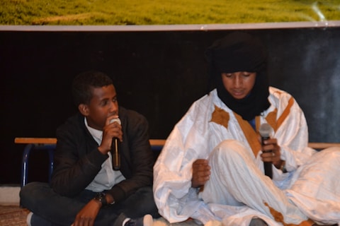Two seated individuals are holding microphones while engaged in a conversation. One individual is dressed in a casual black jacket and blue jeans, while the other is wearing a traditional white and brown robe with a black headscarf.