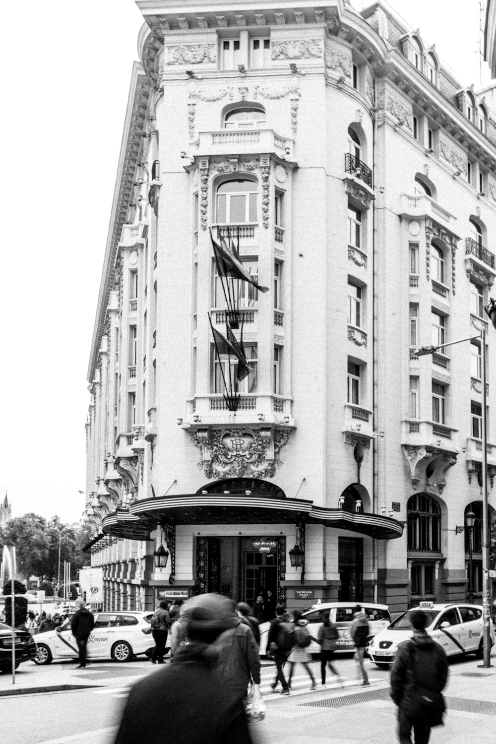 grayscale photo of people and vehicles crossing street near building