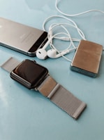 A collection of personal electronic devices laid out on a light blue surface, including a smartphone, a smartwatch with a metal mesh strap, a pair of white earphones, and a small metallic rectangular device.