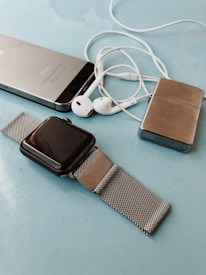 A collection of personal electronic devices laid out on a light blue surface, including a smartphone, a smartwatch with a metal mesh strap, a pair of white earphones, and a small metallic rectangular device.