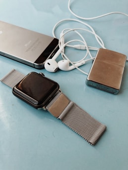 A collection of personal electronic devices laid out on a light blue surface, including a smartphone, a smartwatch with a metal mesh strap, a pair of white earphones, and a small metallic rectangular device.