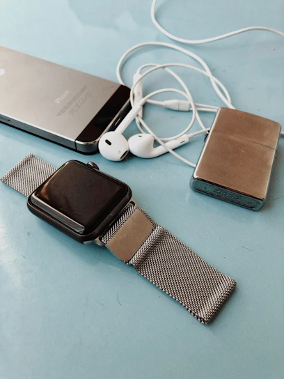 A collection of personal electronic devices laid out on a light blue surface, including a smartphone, a smartwatch with a metal mesh strap, a pair of white earphones, and a small metallic rectangular device.