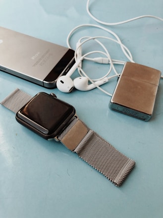 A collection of personal electronic devices laid out on a light blue surface, including a smartphone, a smartwatch with a metal mesh strap, a pair of white earphones, and a small metallic rectangular device.