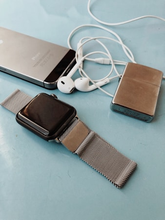 A collection of personal electronic devices laid out on a light blue surface, including a smartphone, a smartwatch with a metal mesh strap, a pair of white earphones, and a small metallic rectangular device.