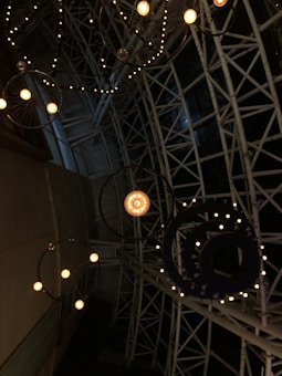 A complex ceiling structure with geometric patterns and decorative elements, including circular frames adorned with small, warm lights. The metal framework creates an intricate lattice against a dark backdrop.