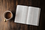 A sunlit Bible open on a wooden table with a steaming cup of tea nearby.