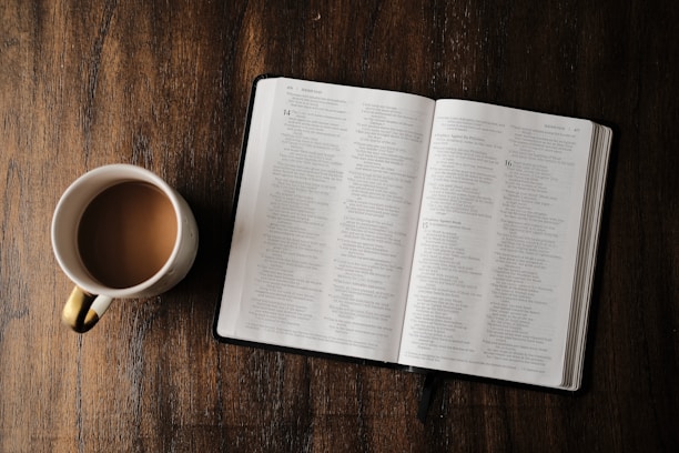 An open Bible resting on a wooden table beside a steaming cup of tea.