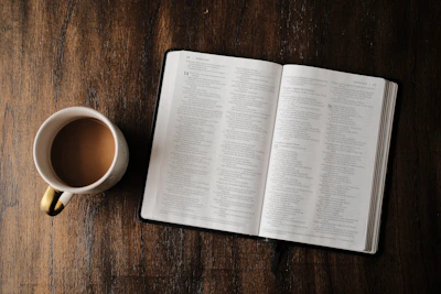 Open Bible resting on worn wooden table next to a steaming cup of coffee.
