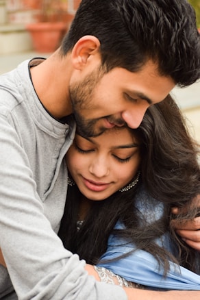 A couple embraces warmly, with their heads closely touching. The woman is smiling gently with her eyes closed while being held by the man, who also has his eyes closed. They both appear content and intimate. The background includes blurred outdoor elements like a plant pot.