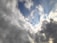 A dynamic sky scene featuring a mix of dark and light clouds with patches of blue sky peeking through. The clouds vary in thickness and texture, with some areas appearing more turbulent while others are softer. Sunlight creates a bright highlight on some clouds, enhancing the contrast and depth.