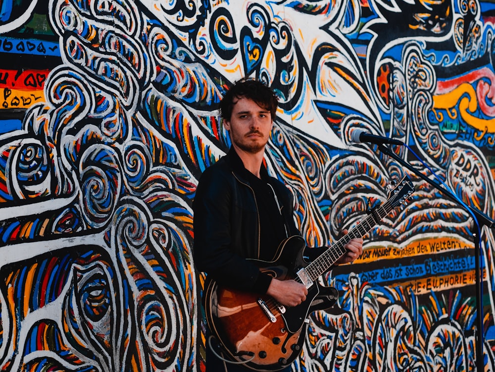 man holding brown guitar standing beside graffiti wall