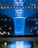 A spectacular indoor waterfall illuminated with blue lighting, surrounded by a geometric glass and steel dome. The scene includes reflections and ambient greenery visible in the background, creating a futuristic and serene atmosphere.
