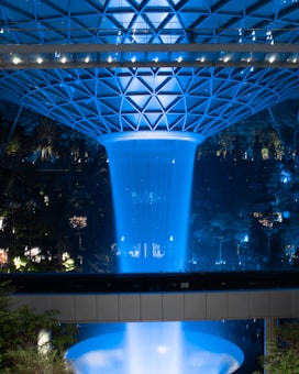 A spectacular indoor waterfall illuminated with blue lighting, surrounded by a geometric glass and steel dome. The scene includes reflections and ambient greenery visible in the background, creating a futuristic and serene atmosphere.