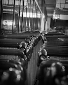A monochrome image of a row of shopping carts lined up in a supermarket or store environment. The carts are tightly packed together, and the image is captured from a low angle, creating a sense of depth. The lighting is dim, with reflections on the metal surfaces, giving the scene an industrial feel.